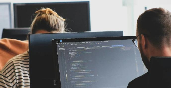 Two people working on computer code at monitors in a bright office workspace