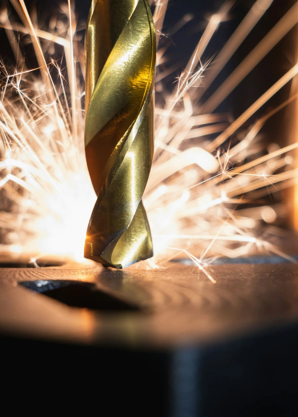 A gold metal drill bit cutting into a dark surface with bright sparks